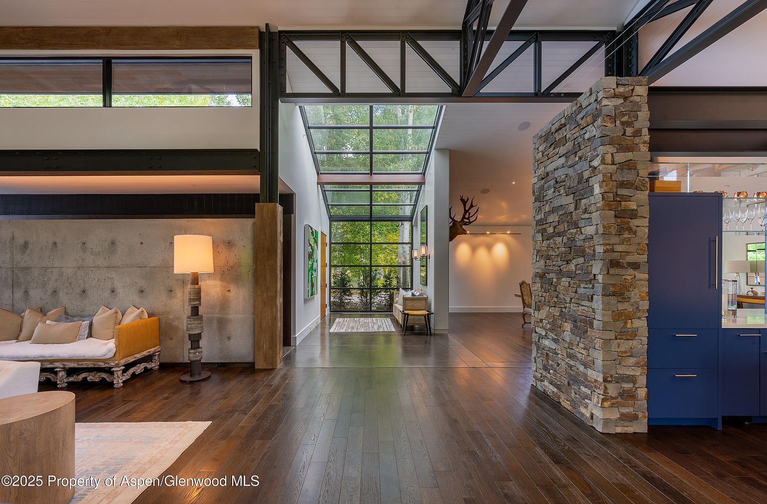 This interior shot showcases a modern living space with hardwood floors and a blend of textures, including concrete and stone accents. A large window wall provides ample natural light, highlighting the open floor plan and contemporary design. The space is furnished with comfortable seating and unique lighting fixtures, creating an inviting atmosphere.
