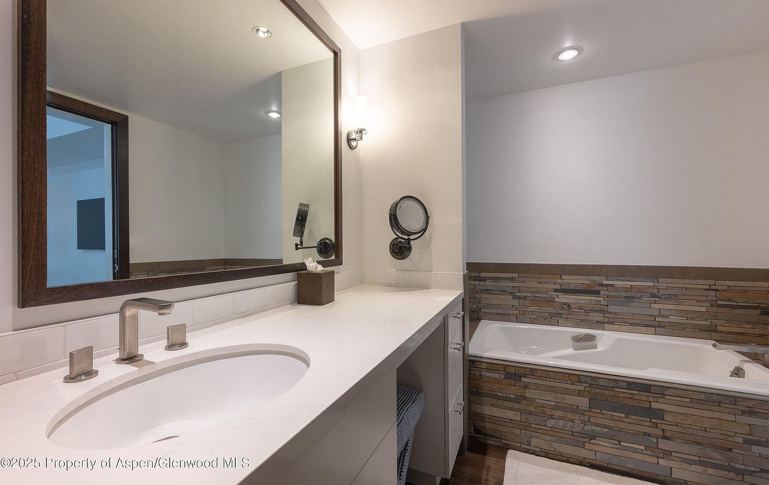 This is a well-lit primary bathroom featuring a large mirror with a dark wood frame above a white countertop with an integrated sink and modern faucet. A bathtub with stone accents is visible in the background, adding a touch of luxury and natural texture to the space. The overall impression is clean, modern, and inviting.