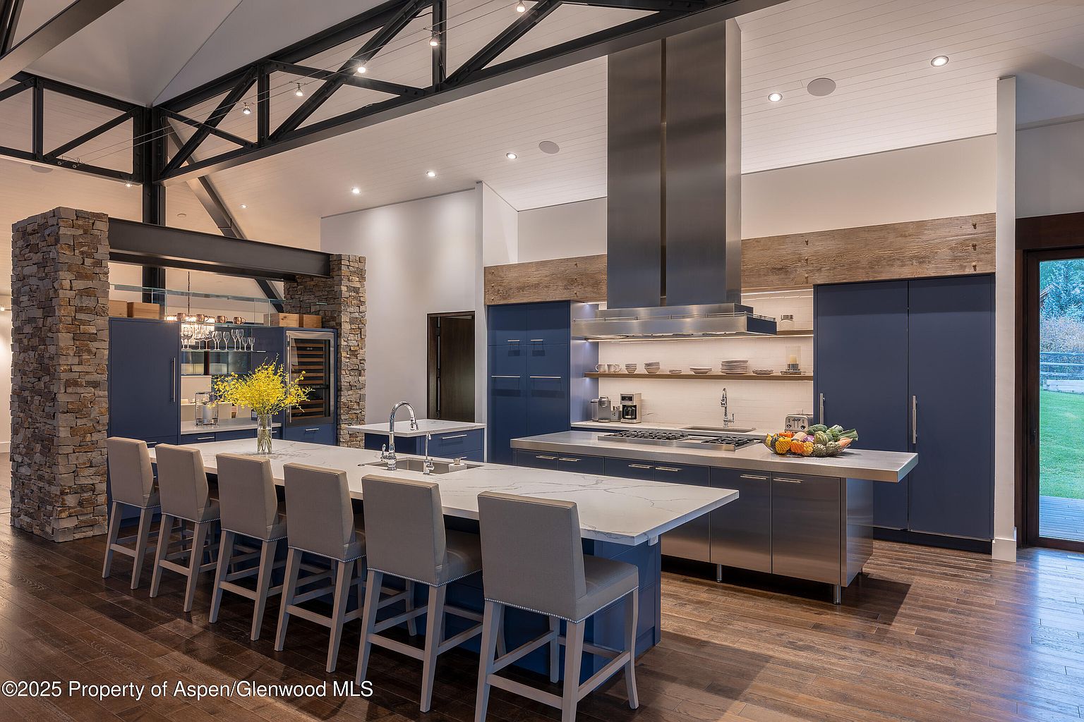 This is a modern kitchen with dark blue cabinetry, stainless steel appliances, and a large island with a white countertop. The kitchen features a unique overhead beam structure and a stainless steel range hood. The overall style is contemporary with rustic touches.