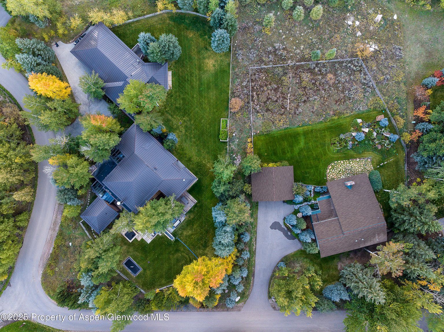 This aerial view showcases a luxurious residential property with multiple structures, meticulously landscaped grounds, and mature trees displaying vibrant fall colors. The homes feature dark-colored roofs and are surrounded by lush lawns, gardens, and natural vegetation, creating a serene and private setting. A winding driveway connects the buildings, enhancing the estate's overall appeal.