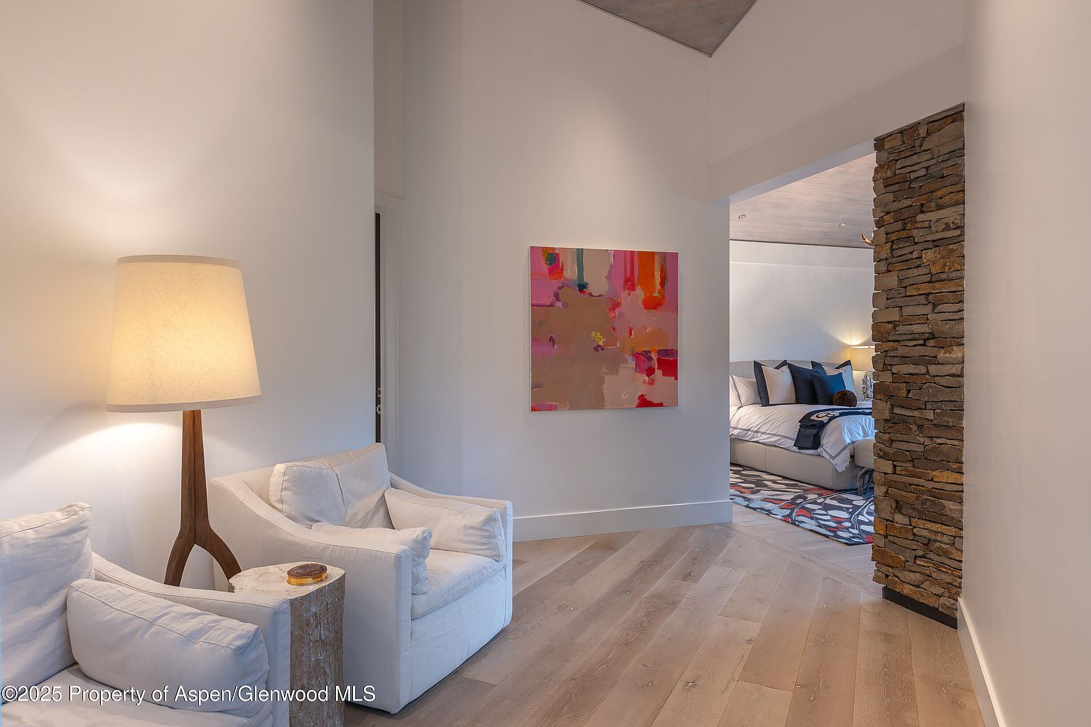 This interior shot showcases a bright hallway leading into a bedroom. The hallway features light wood flooring, white walls adorned with modern art, and a stone accent wall framing the bedroom entrance. The bedroom is visible in the background, featuring a bed with blue accents and a patterned rug, creating a luxurious and inviting atmosphere.