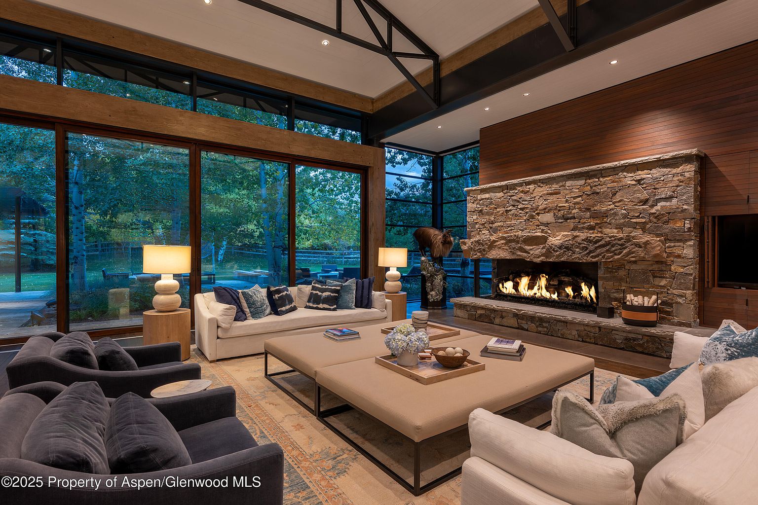 This is a luxurious living room featuring a stone fireplace, large windows with views of the outdoors, and comfortable seating arrangements. The room is well-lit with lamps and natural light, creating a warm and inviting atmosphere. The neutral color palette and natural materials contribute to a sophisticated and relaxing ambiance.