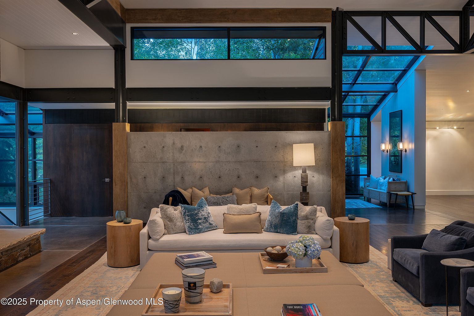 This is an interior shot of a modern living room featuring a neutral-toned sofa adorned with decorative pillows, complemented by a large ottoman serving as a coffee table. The room is characterized by its contemporary design, incorporating elements like exposed steel beams, a concrete accent wall, and large windows that provide natural light. The space is furnished with wooden side tables and a comfortable armchair, creating a cozy yet sophisticated atmosphere.