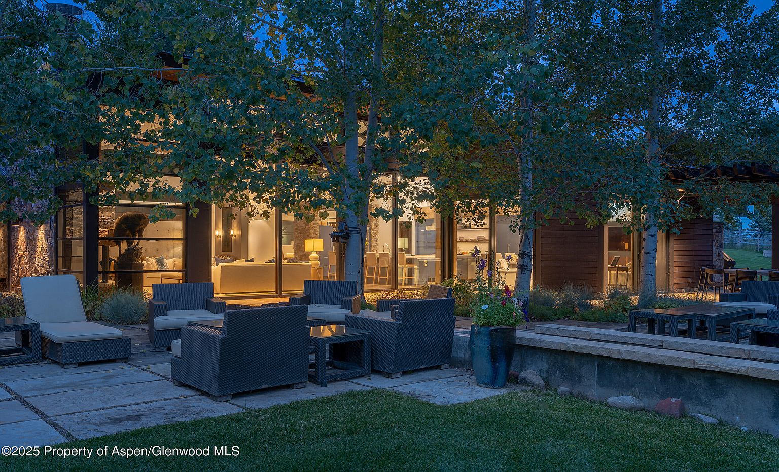 This image showcases an inviting outdoor patio area, featuring comfortable wicker seating arrangements around glass-topped tables. The patio is paved with stone tiles and is surrounded by lush greenery, including trees and manicured grass. Large glass windows offer a glimpse into the home's interior, creating a seamless transition between indoor and outdoor living spaces.