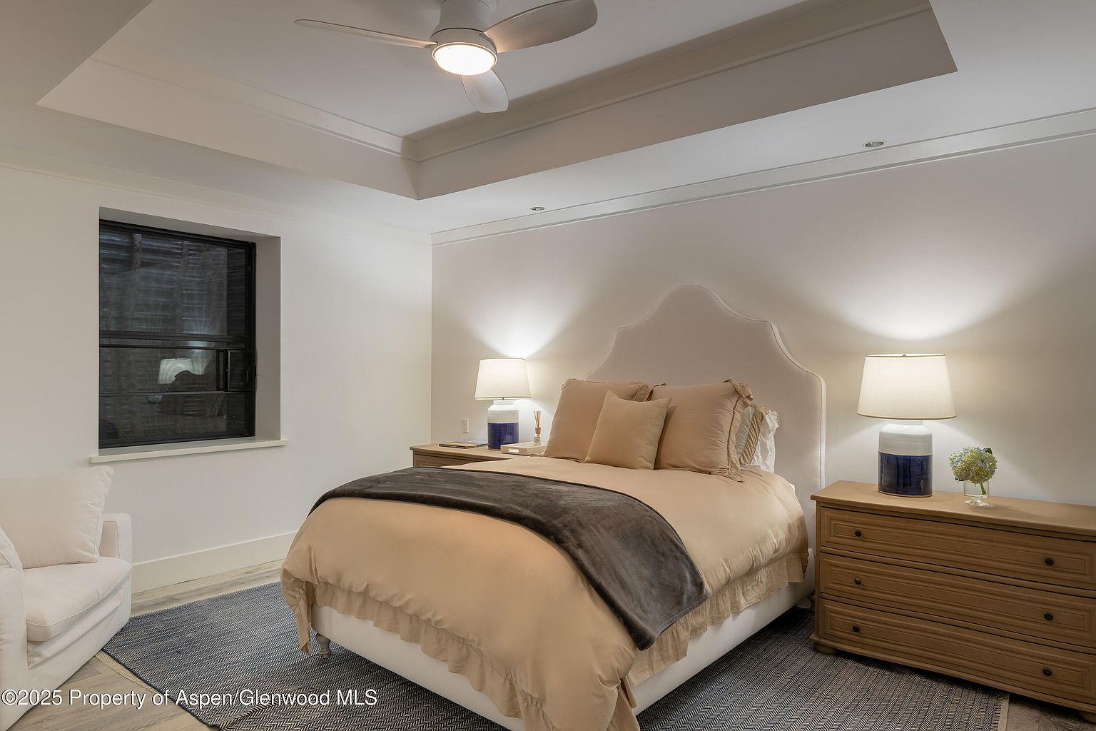 This is a well-lit primary bedroom featuring a neutral color palette. The room includes a bed with a decorative headboard, bedside tables with lamps, and a dresser. A ceiling fan and a window provide additional light and ventilation, creating a serene and inviting atmosphere.