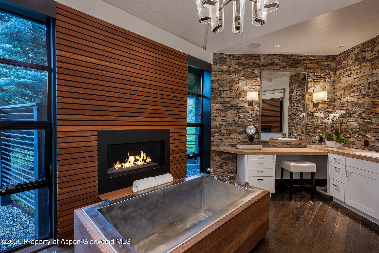This is a luxurious primary bathroom featuring a modern design with a blend of natural elements. The focal point is a unique metal bathtub encased in wood, positioned next to a fireplace with a wood-paneled wall. A vanity area with stone walls, a mirror, and white cabinetry adds to the spa-like ambiance, complemented by dark wood flooring and a contemporary chandelier.