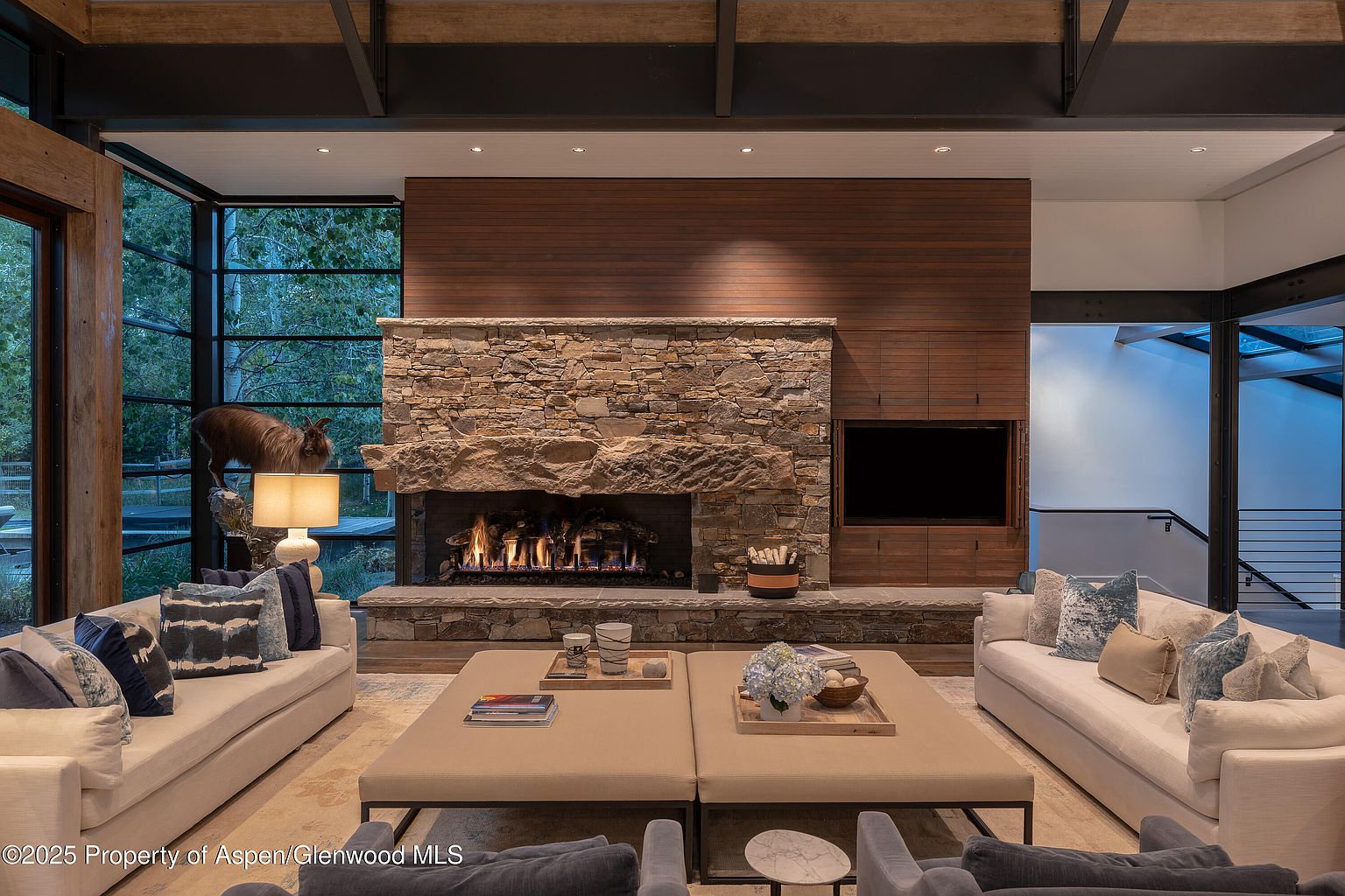 This is an interior shot of a luxurious living room featuring a stone fireplace with a wooden accent wall above. Two light-colored sofas face each other, flanking a pair of large square ottomans serving as coffee tables. The room is well-lit, with a large window on the left offering a view of the outdoors, and the overall design exudes a warm and inviting atmosphere.
