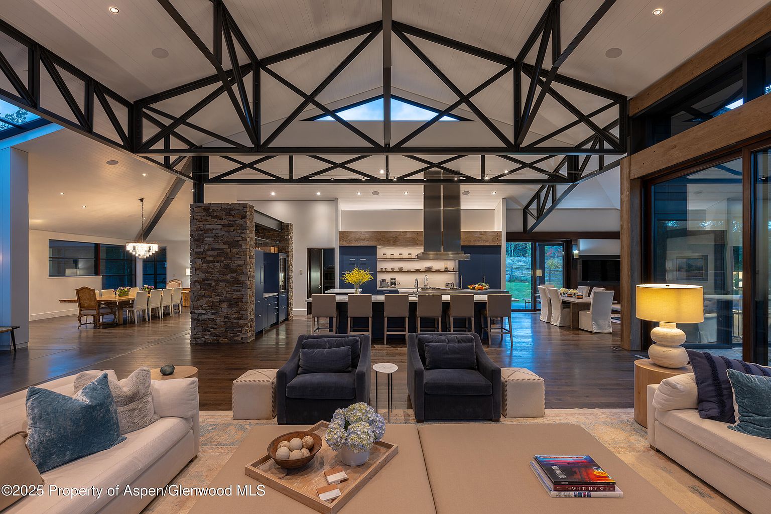 This expansive living room showcases an open-concept design with dark hardwood floors, a striking black beam ceiling structure, and a stone-clad feature wall. The space is furnished with comfortable sofas, armchairs, and a large kitchen island with bar seating in the background, creating an inviting atmosphere for relaxation and entertainment. Natural light floods the room through large windows, enhancing the sense of spaciousness and luxury.