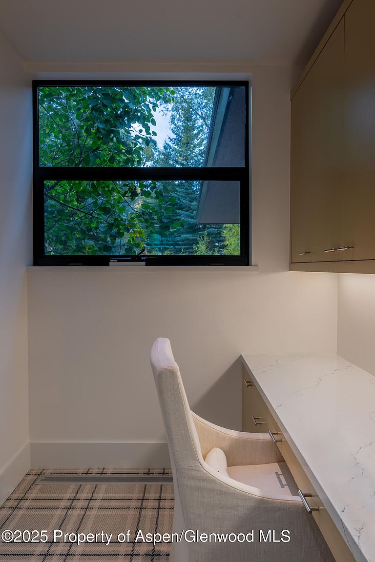 This is an interior shot of a small office or study area. The room features a built-in desk with a light-colored countertop, a comfortable chair, and a window that provides natural light and a view of the trees outside. The overall impression is a clean and functional workspace.