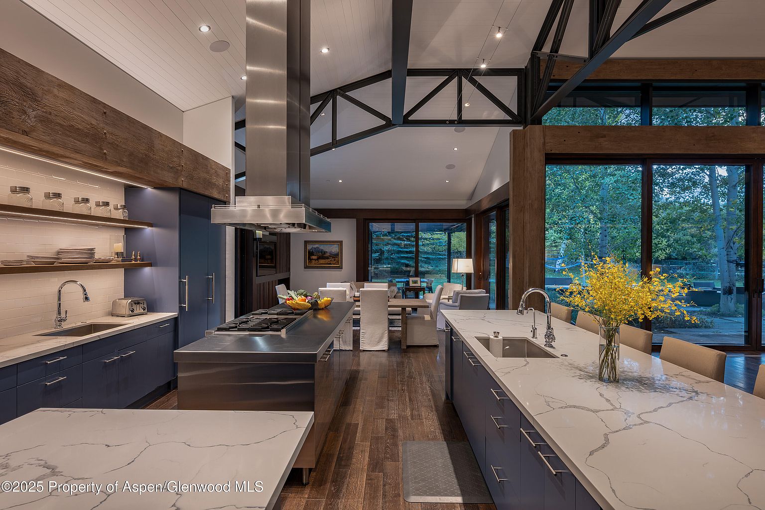 This is a view of a modern kitchen with dark blue cabinets and a large island with a white countertop. The kitchen features stainless steel appliances and a large window with a view of the outdoors. The overall style is contemporary and luxurious, with a focus on natural light and open space.