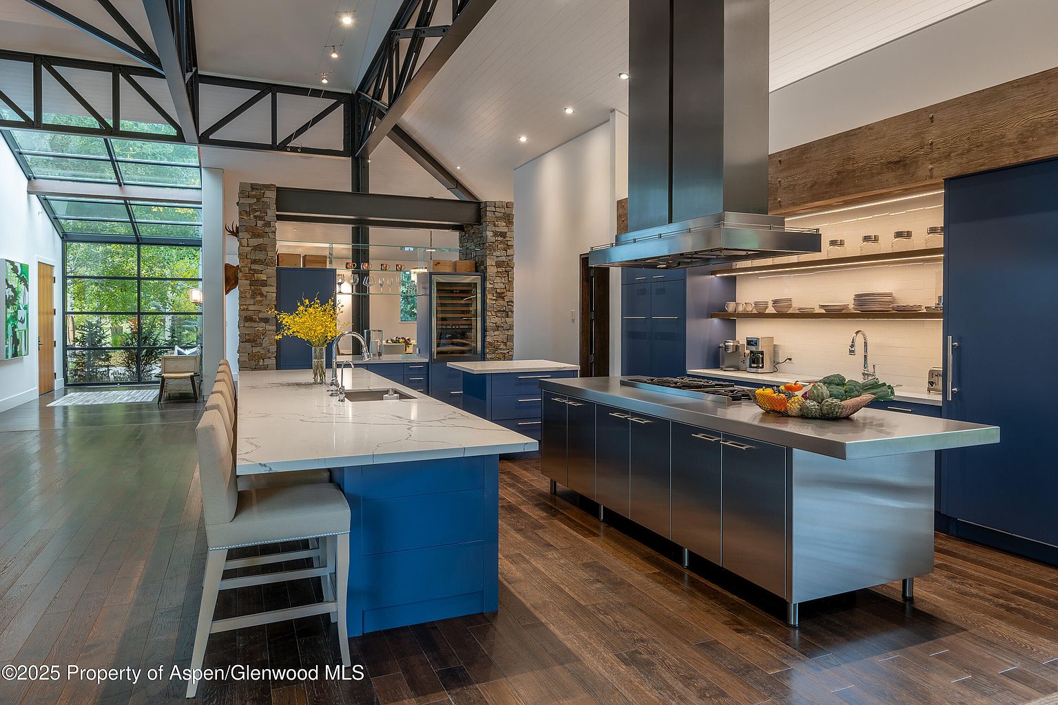 This is a modern kitchen with a large island featuring a white countertop and blue cabinetry. The kitchen also features stainless steel appliances, including a range hood, and a second island with a stainless steel countertop. The space is well-lit and has a contemporary design with a mix of materials, including wood flooring and stone accents.