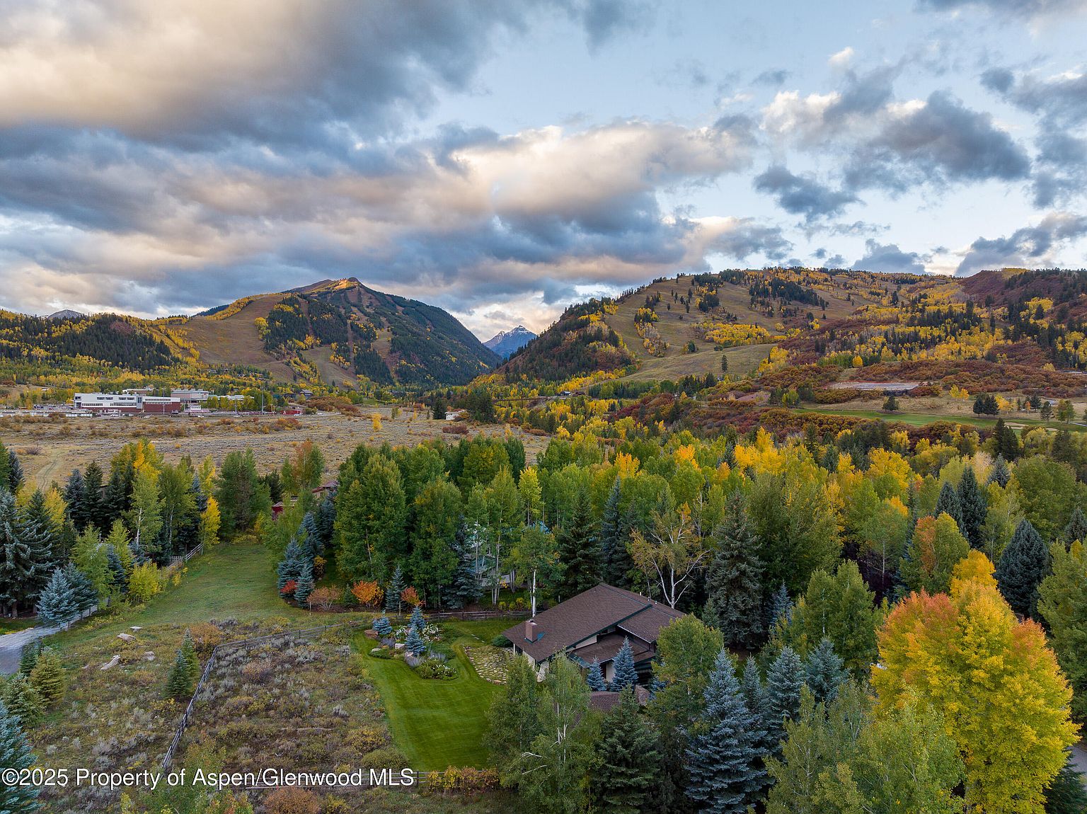 This aerial shot showcases a beautiful property nestled amidst a vibrant landscape of fall foliage and rolling hills. The house, featuring a brown roof, is surrounded by lush greenery and mature trees, creating a sense of privacy and tranquility. The overall impression is one of a serene and luxurious mountain retreat.