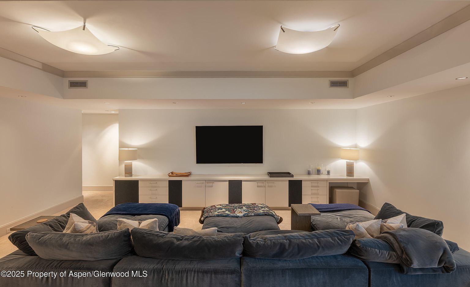 This is an interior shot of a living room featuring a large, plush sectional sofa in the foreground. The room is well-lit with modern ceiling fixtures and lamps on a long console table against the wall, where a flat-screen TV is mounted. The style is contemporary and comfortable, suggesting a relaxing entertainment space.