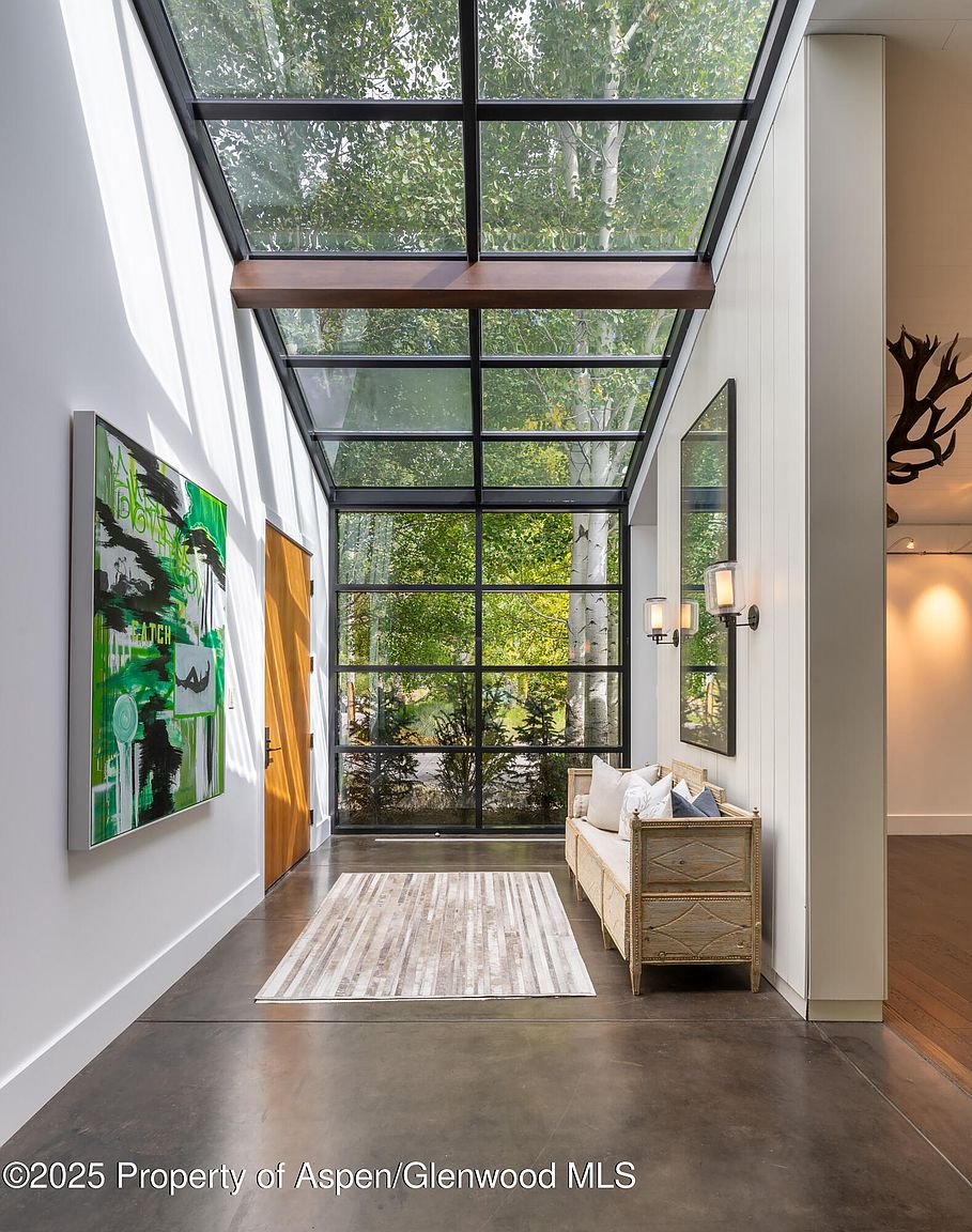 This interior shot showcases a modern hallway with a striking glass ceiling, allowing natural light to flood the space. A contemporary painting adorns the left wall, while a stylish bench sits against the right wall, complemented by a mirror and sconces. The polished concrete floor and minimalist design create a sophisticated and inviting atmosphere.