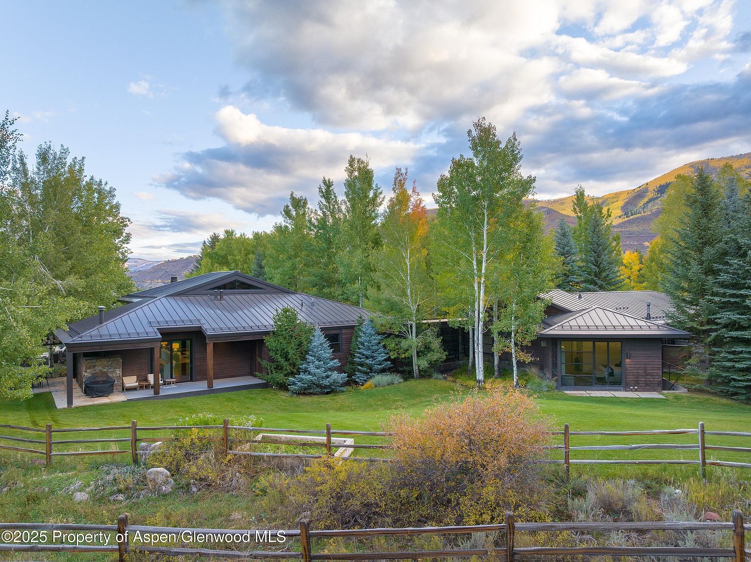 This image showcases the front exterior of a beautiful home with a dark metal roof and wood siding. The property features a well-manicured lawn, mature trees, and a rustic wooden fence. The architecture blends seamlessly with the natural surroundings, creating a serene and inviting atmosphere.