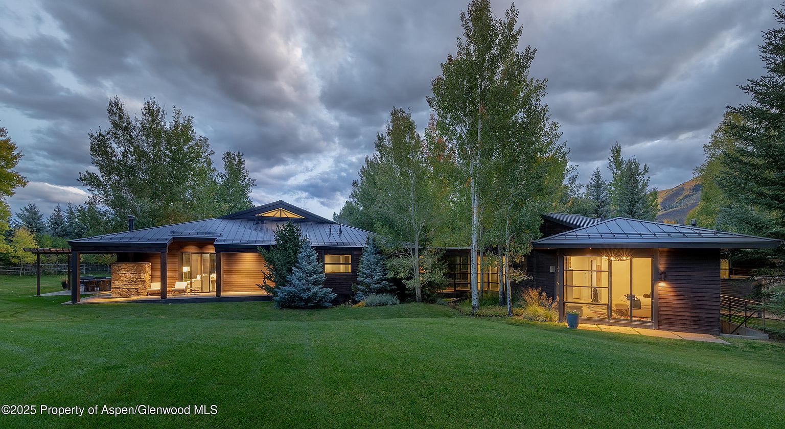 This is a front exterior view of a modern home with a dark metal roof and dark wood siding. The house is surrounded by a lush green lawn and mature trees, creating a serene and private setting. Large windows provide ample natural light and offer views of the surrounding landscape, enhancing the property's appeal.
