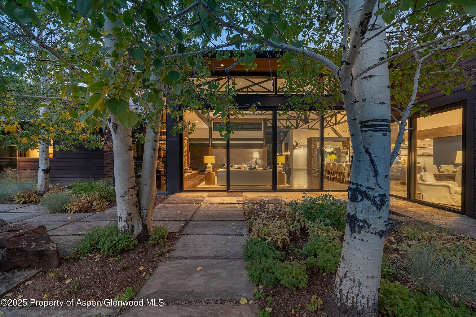 This image showcases the front exterior of a modern home, emphasizing its architectural design and landscaping. Large glass windows and doors provide a view into the living spaces, while the stone pathway and birch trees create an inviting entrance. The overall impression is one of contemporary elegance and natural integration.