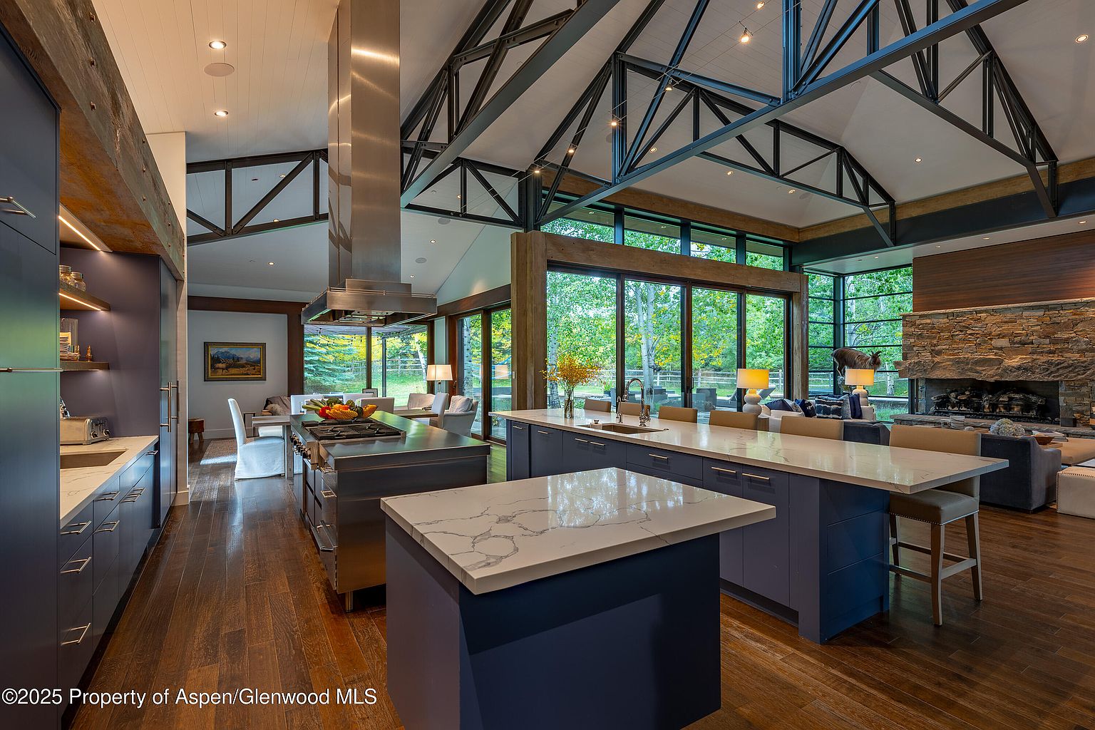 This is a spacious, modern kitchen with dark wood floors and a high, vaulted ceiling with exposed beams. The kitchen features two large islands with white marble countertops and dark blue cabinetry. Large windows offer views of the surrounding greenery, and the kitchen seamlessly transitions into a living area with a stone fireplace.