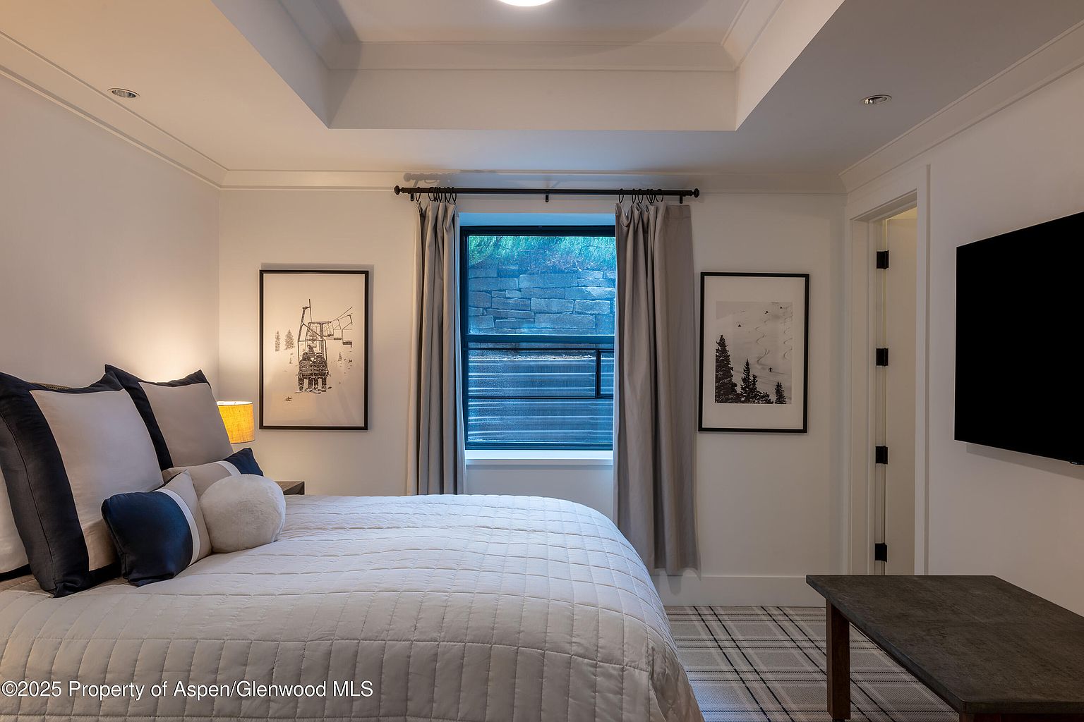 This is a well-lit primary bedroom featuring a bed with plush pillows and a quilted comforter. The room has a neutral color palette with white walls and framed artwork, complemented by a window with gray curtains. A dark wood table sits near a wall-mounted television, adding a touch of modern contrast.