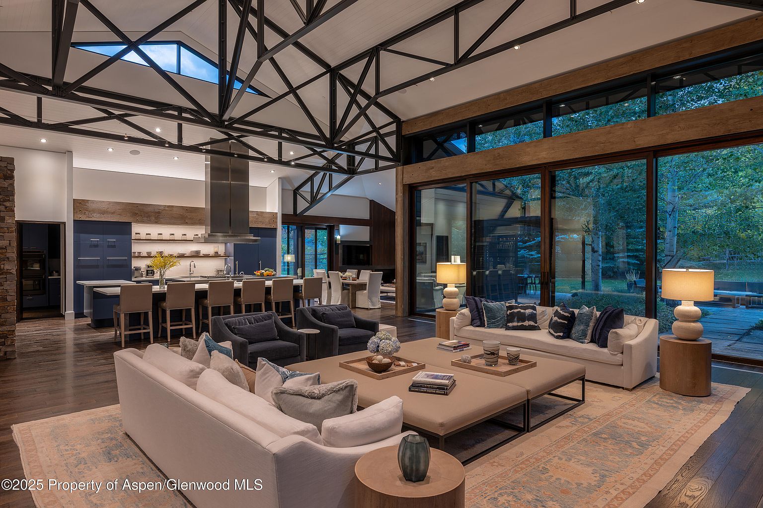 This is a spacious and luxurious living room featuring an open-concept design that seamlessly integrates with the kitchen. The room is furnished with plush white sofas, navy armchairs, and a large ottoman coffee table, all arranged on a patterned rug. Floor-to-ceiling windows offer views of the outdoors, while the exposed beam ceiling adds architectural interest and a sense of grandeur.