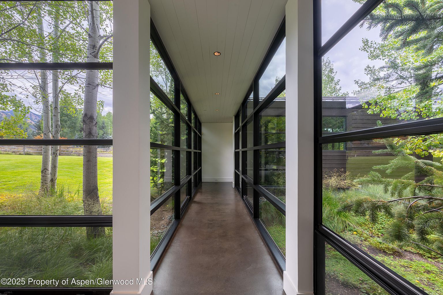 This image showcases a modern hallway with floor-to-ceiling windows on both sides, offering expansive views of the surrounding landscape. The hallway features a sleek, minimalist design with white pillars and a dark-toned floor, creating a bright and airy atmosphere. The architectural style emphasizes clean lines and a seamless connection between the interior and exterior spaces.
