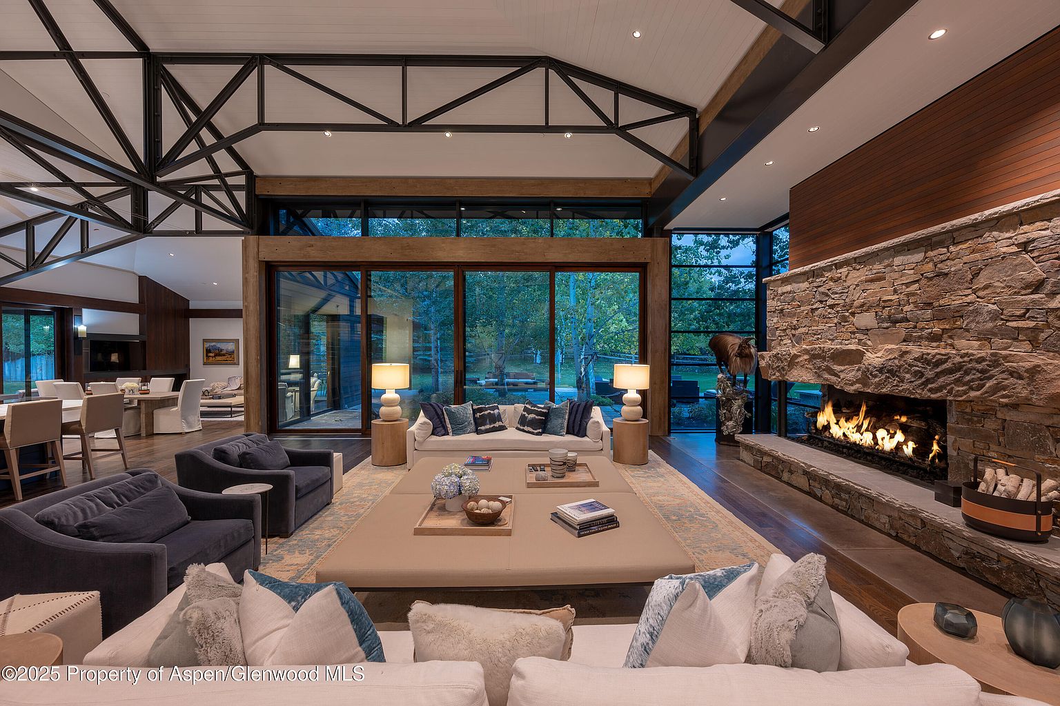 This is a spacious living room featuring a stone fireplace, large windows with views of the outdoors, and comfortable seating arrangements. The room has a high ceiling with exposed beams, creating a modern yet rustic atmosphere. The hardwood floors and neutral color palette add to the room's warmth and inviting feel.
