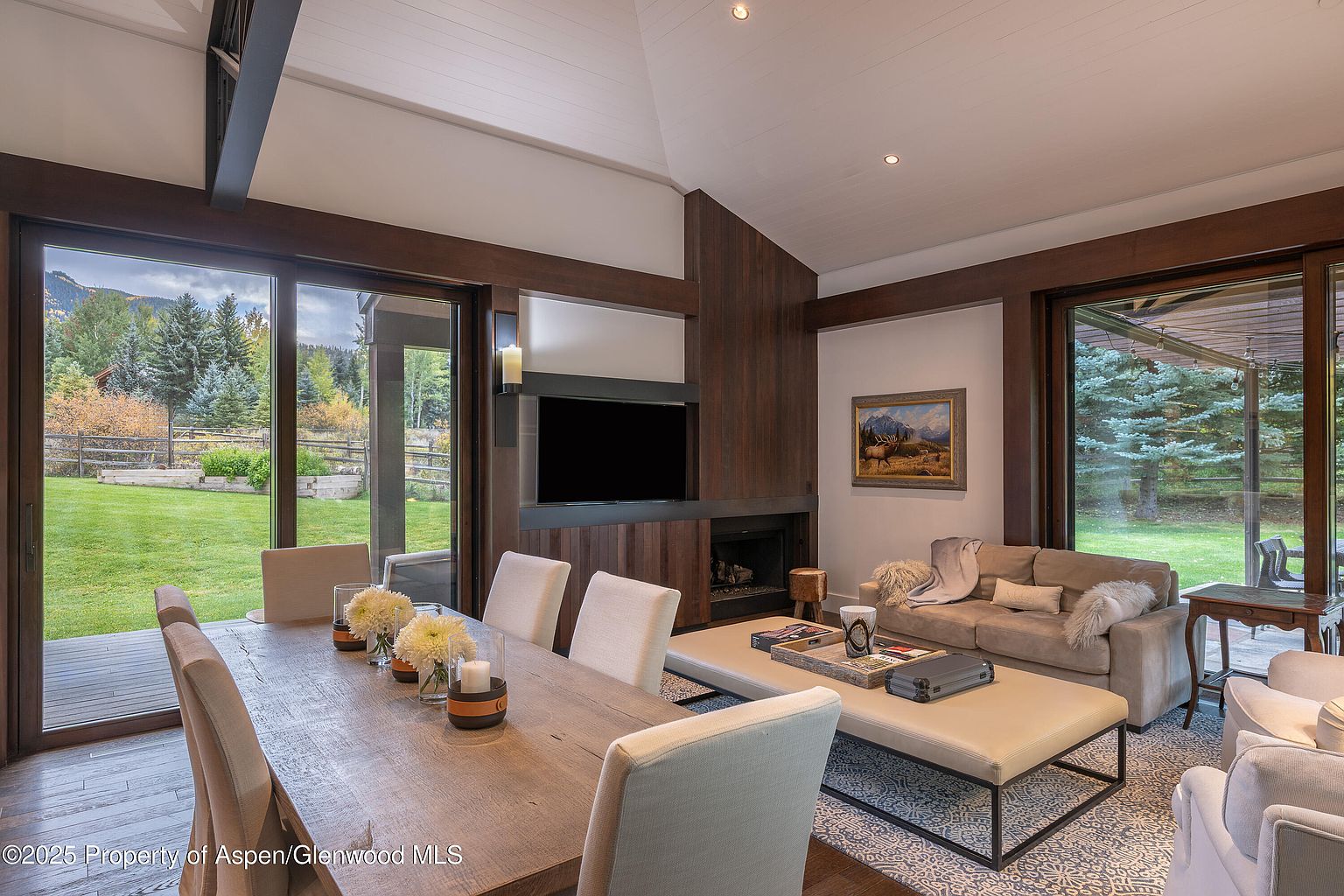 This is an interior shot of a combined living and dining area. The space features a long wooden dining table with upholstered chairs, a comfortable sofa with a large ottoman, and a modern fireplace with a mounted television. Large windows offer views of the outdoor landscape, creating a bright and inviting atmosphere.