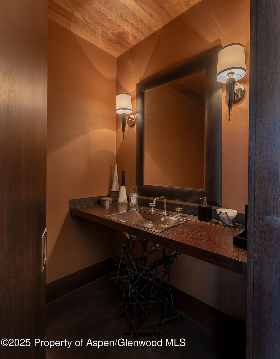 This is a cozy guest bathroom featuring warm, earthy tones and rustic design elements. The vanity has a unique metal base and a dark wood countertop with a vessel sink. A framed mirror is flanked by sconce lighting, and the walls are painted in a rich, terracotta hue, creating a warm and inviting atmosphere.