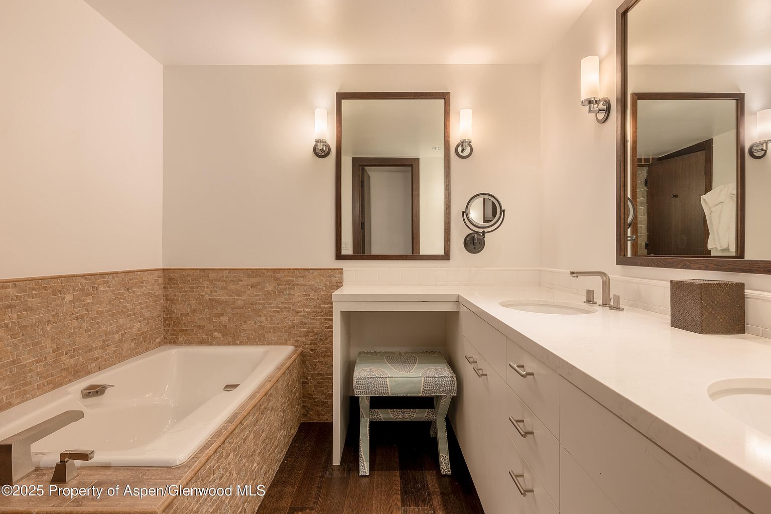 This is a well-lit primary bathroom featuring a double vanity with white countertops and cabinetry. Two framed mirrors hang above the sinks, each flanked by sconce lighting. A bathtub with tiled surround is visible to the left, and a small upholstered stool sits beneath a vanity area, adding a touch of elegance and functionality to the space.