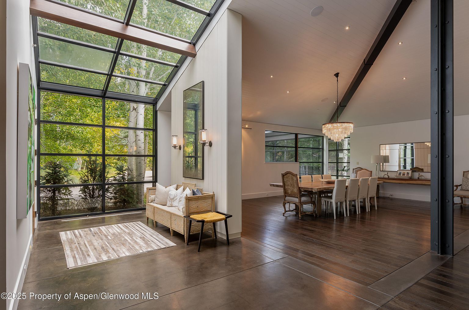 This interior shot showcases a combined entryway and dining area, featuring a striking glass-enclosed space with views of lush greenery. The dining area boasts a long wooden table surrounded by elegant chairs and illuminated by a modern chandelier. The flooring transitions from concrete in the entryway to hardwood in the dining area, adding visual interest and defining the spaces.