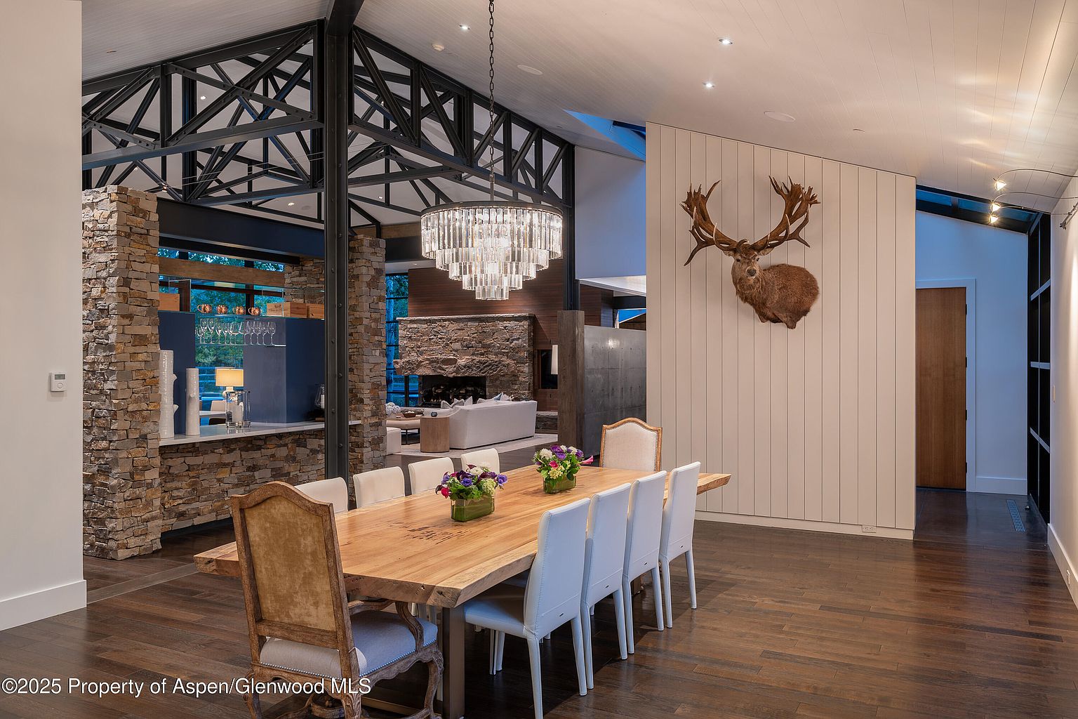 This interior shot showcases a dining room with a large wooden table surrounded by white chairs and two upholstered chairs at the ends. A striking chandelier hangs above the table, and a mounted deer head adorns the wall. The room features hardwood floors and a stone accent wall, creating a blend of rustic and modern design elements.