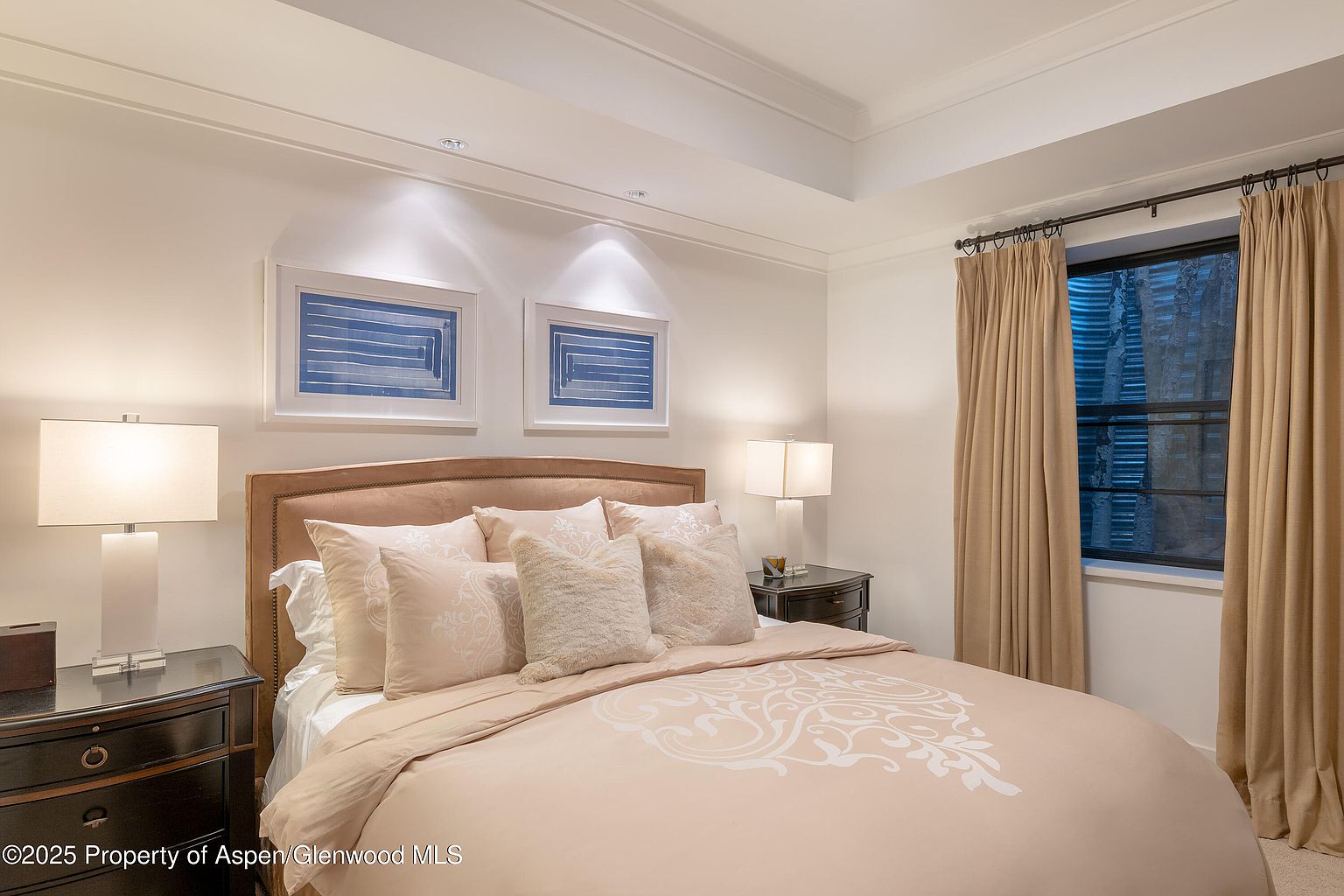 This is a well-lit primary bedroom featuring a bed with a beige headboard and matching bedding with a subtle pattern. Two framed blue artworks hang above the headboard, flanked by bedside tables with lamps. A window with beige curtains provides natural light, creating a cozy and inviting atmosphere.