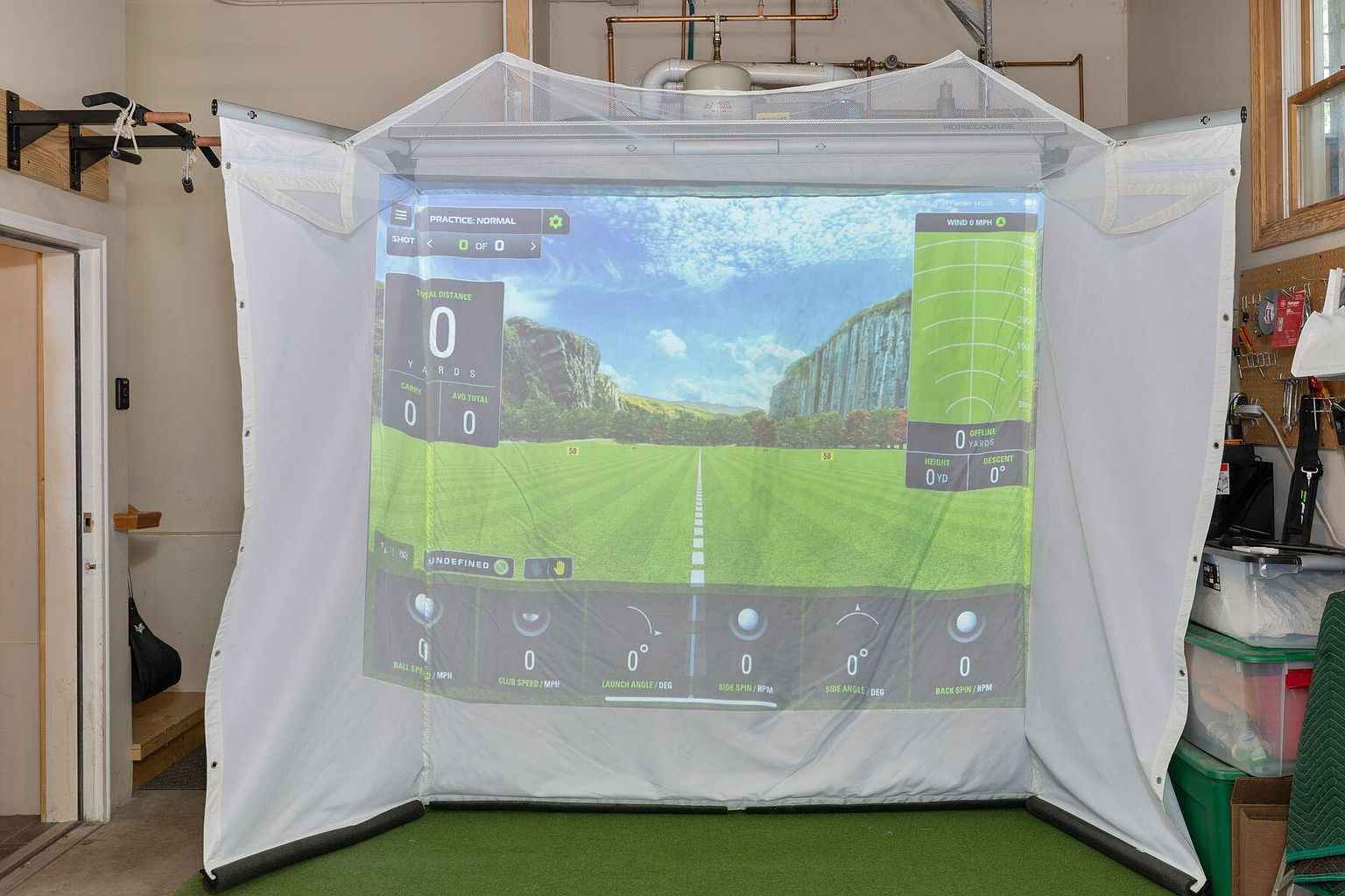 This image showcases a home golf simulator setup, featuring a large projection screen displaying a virtual golf course. The simulator is enclosed by a white fabric enclosure, creating a dedicated practice space. The setup includes artificial turf and displays various golf metrics on the screen, suggesting a high-tech and immersive training environment.
