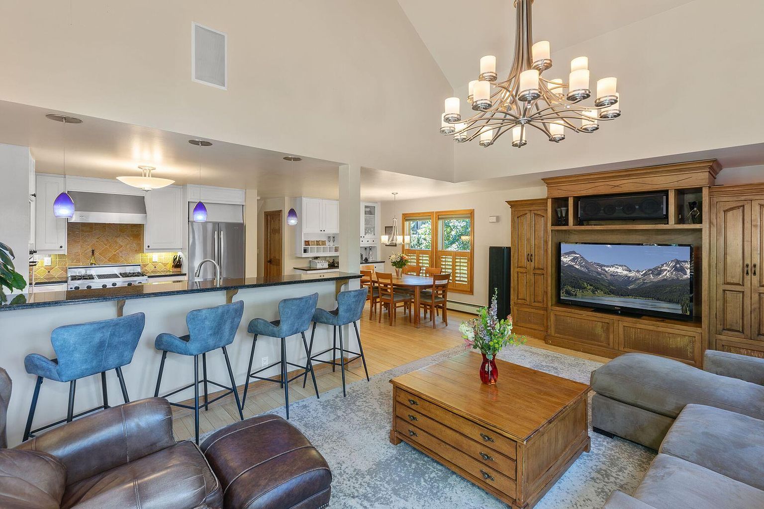 This is an interior shot of a combined living room, dining area, and kitchen. The living room features a large wooden entertainment center with a TV, a coffee table, and comfortable seating. The kitchen has a breakfast bar with blue bar stools, stainless steel appliances, and white cabinetry. The dining area has a wooden table and chairs, creating a warm and inviting atmosphere.