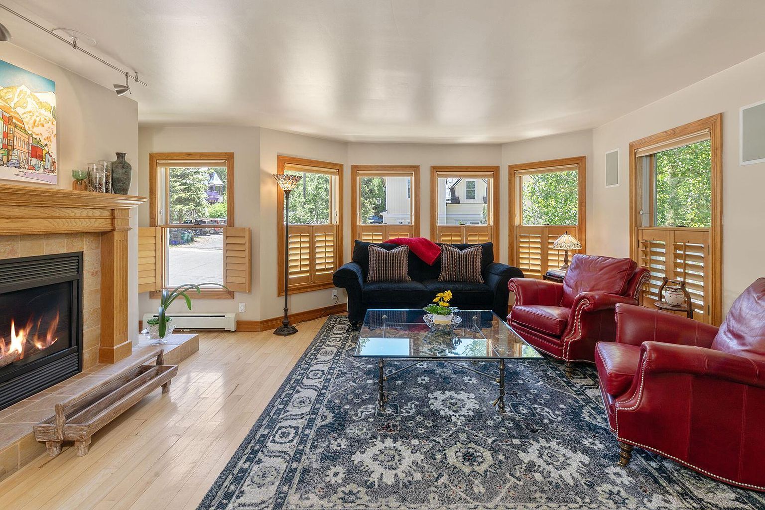 This is a warm and inviting living room featuring a fireplace with a wooden mantel, a black sofa with decorative pillows, and two red leather armchairs. A glass coffee table sits atop a patterned area rug, and natural light streams in through multiple windows with wooden shutters. The overall impression is one of comfort and classic elegance.