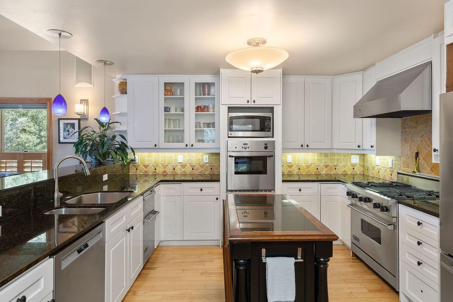 This is a well-lit kitchen featuring white cabinetry, stainless steel appliances, and a dark granite countertop. A kitchen island with a dark wood base and a glass top sits in the center of the room. The backsplash is a patterned tile, and the flooring is light-colored hardwood, creating a warm and inviting atmosphere.