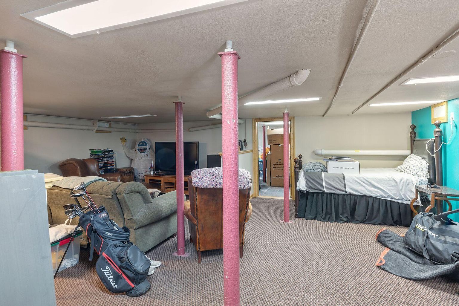 This is a basement interior featuring multiple support columns painted in a distinctive pink color. The space includes a bed, seating area with a golf bag, and a television, suggesting a multi-purpose recreational area. The carpeted floor and drop ceiling contribute to a finished but somewhat cluttered appearance.