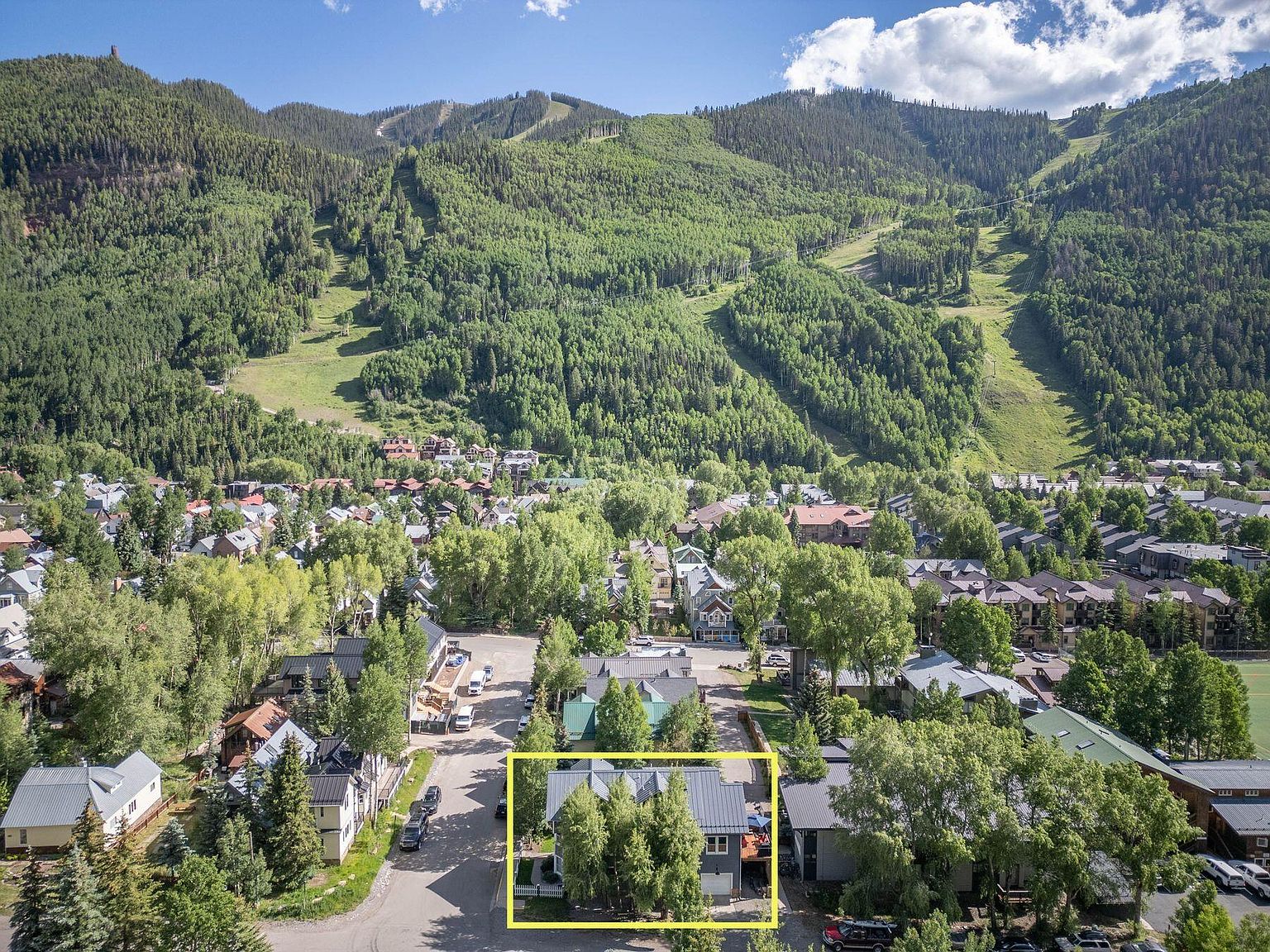 This aerial shot showcases a charming home nestled in a lush, mountainous landscape. The property, outlined in yellow, features a gray exterior, a metal roof, and mature trees providing privacy. The surrounding neighborhood is quaint, with a mix of residential buildings and green spaces, all set against a backdrop of forested hills and a partly cloudy sky.