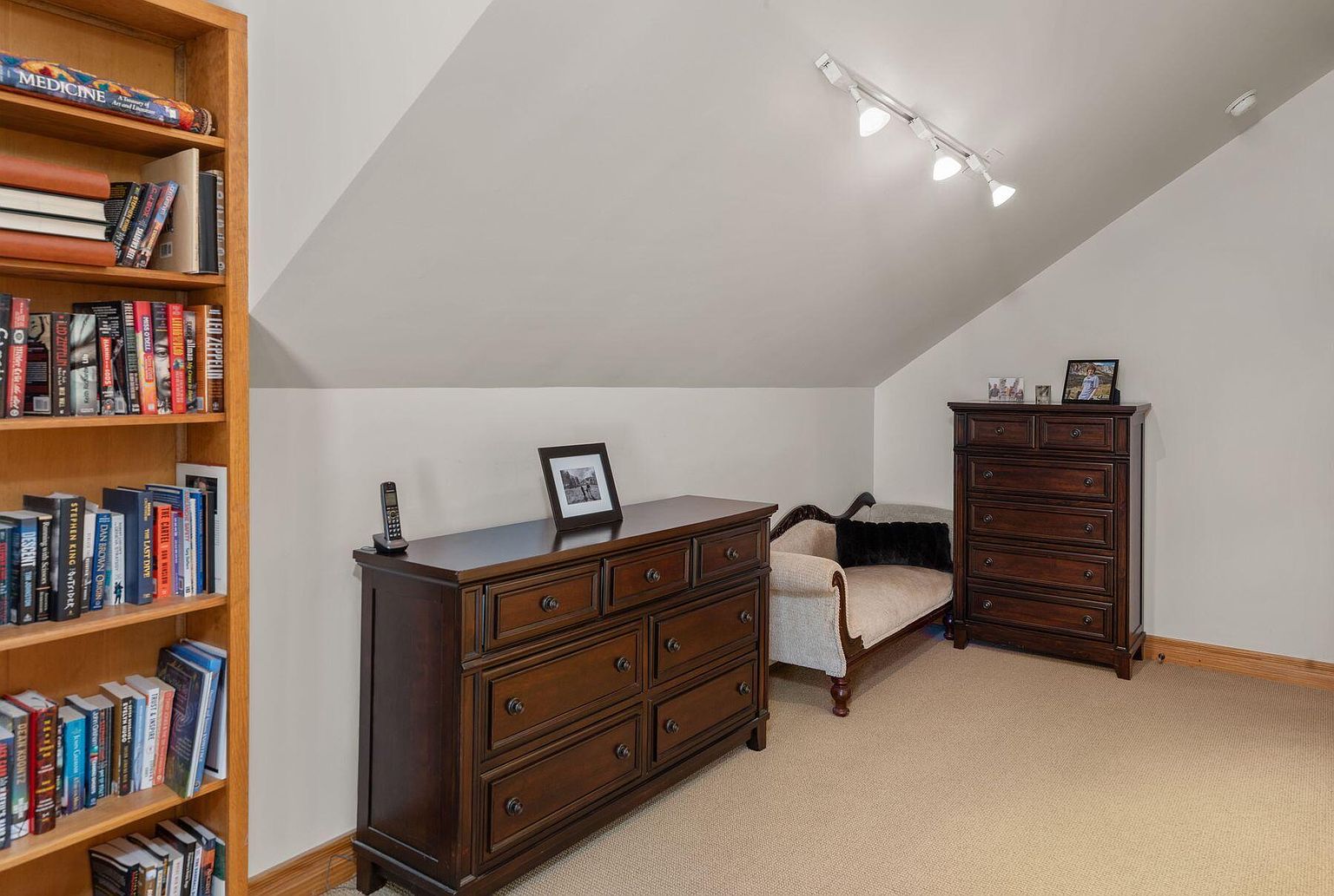 This is an interior shot of a bedroom featuring a bookshelf filled with books, a dark wood dresser with a framed picture and a phone on top, and a chaise lounge with a black pillow. The room has an angled ceiling with track lighting and is carpeted, creating a cozy and inviting atmosphere.