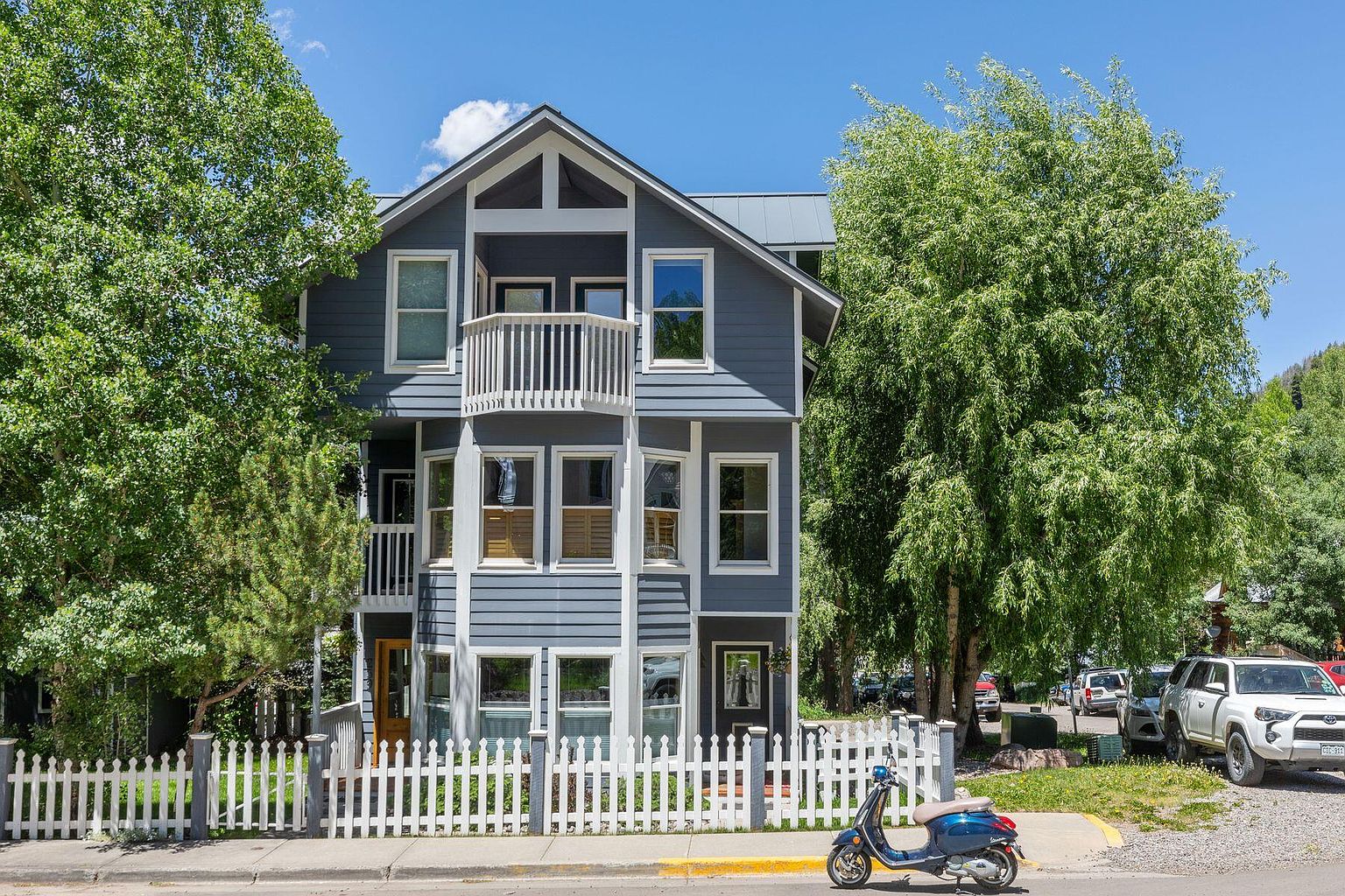 This is a front view of a multi-story house with a gray exterior and white trim. The house features a small balcony on the second floor and a white picket fence surrounding the front yard. A blue scooter is parked on the street in front of the house, adding a touch of character.
