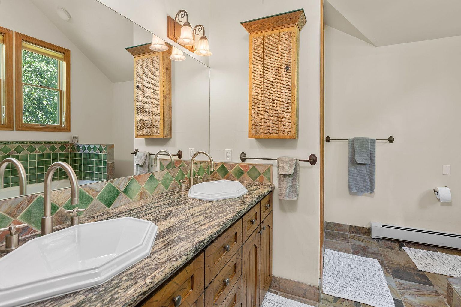 This is a bathroom featuring a double vanity with a granite countertop and two white sinks. The backsplash is tiled with a green and brown diamond pattern. Above the vanity is a large mirror with a wooden-framed light fixture and two wooden cabinets. The floor is tiled with brown slate and there is a white bath mat. The overall style is rustic and cozy.