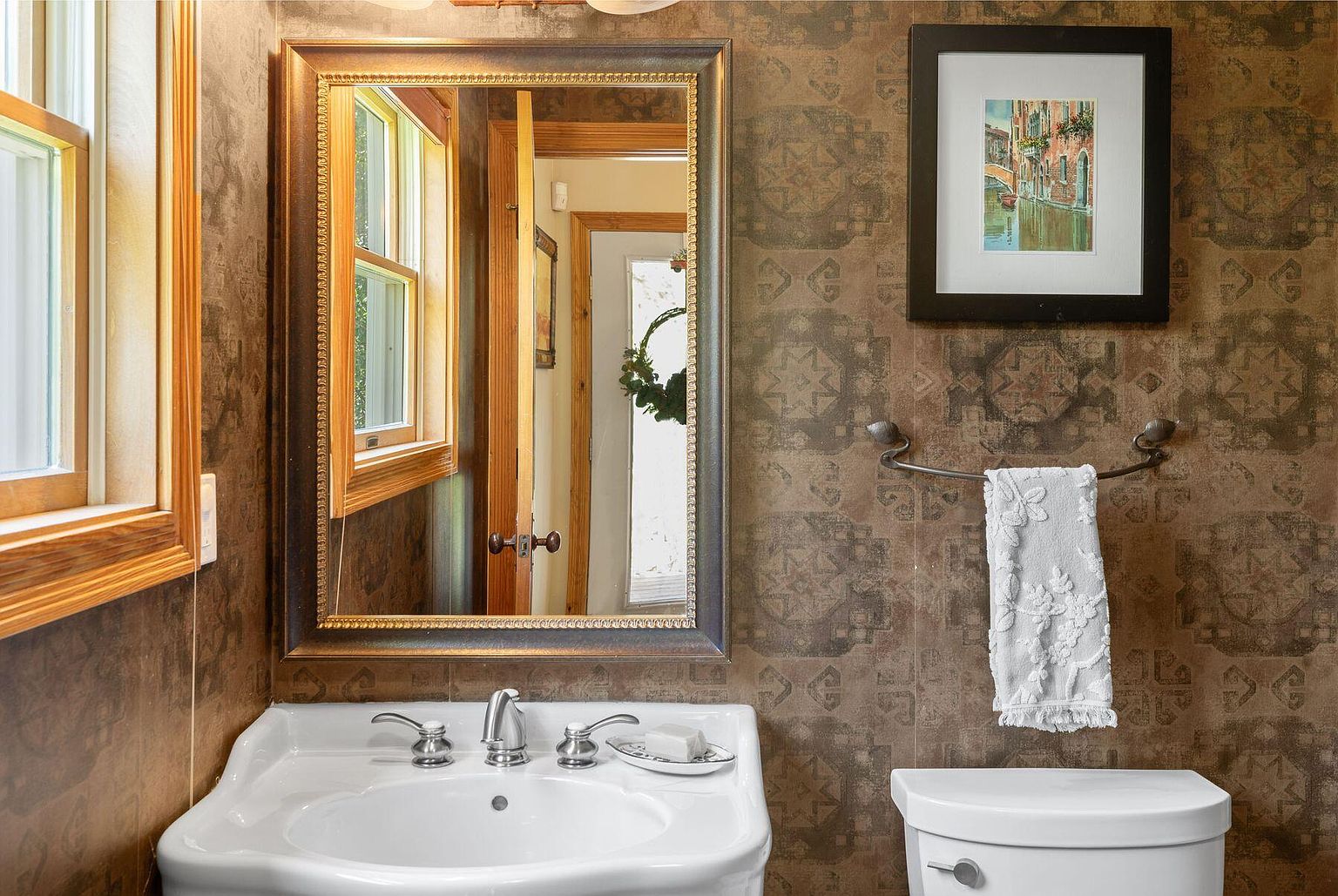This bathroom features a pedestal sink with chrome fixtures, a framed mirror, and a framed picture above a towel rack. The walls are covered in patterned wallpaper, and a toilet is visible to the right. The overall style is traditional and cozy.