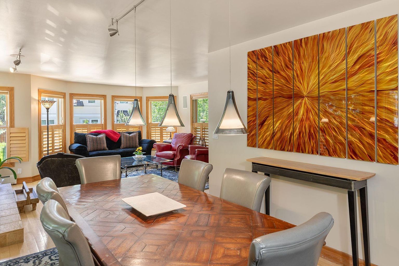 This interior shot showcases a dining room with a large wooden table surrounded by leather chairs. A striking piece of abstract art hangs on the wall, complemented by modern pendant lighting. The room flows into a living area with comfortable seating, creating an inviting and stylish atmosphere.