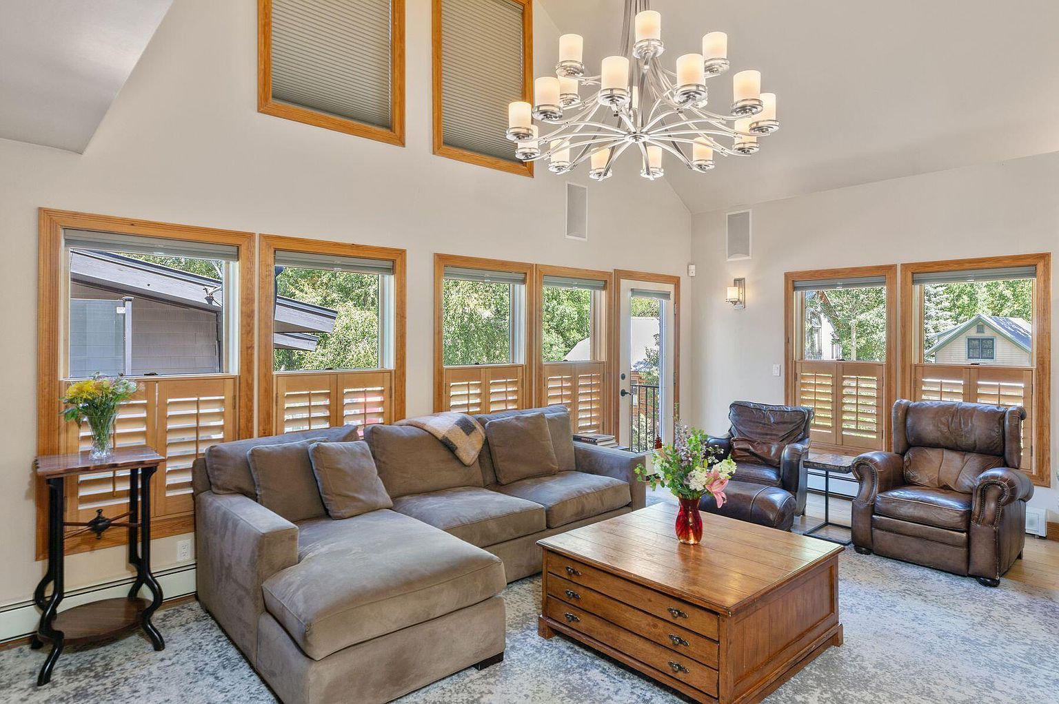 This is a well-lit living room featuring a large sectional sofa, two leather armchairs, and a wooden coffee table. The room has multiple windows with wooden frames and shutters, allowing natural light to fill the space. A modern chandelier hangs from the ceiling, adding a touch of elegance to the room.