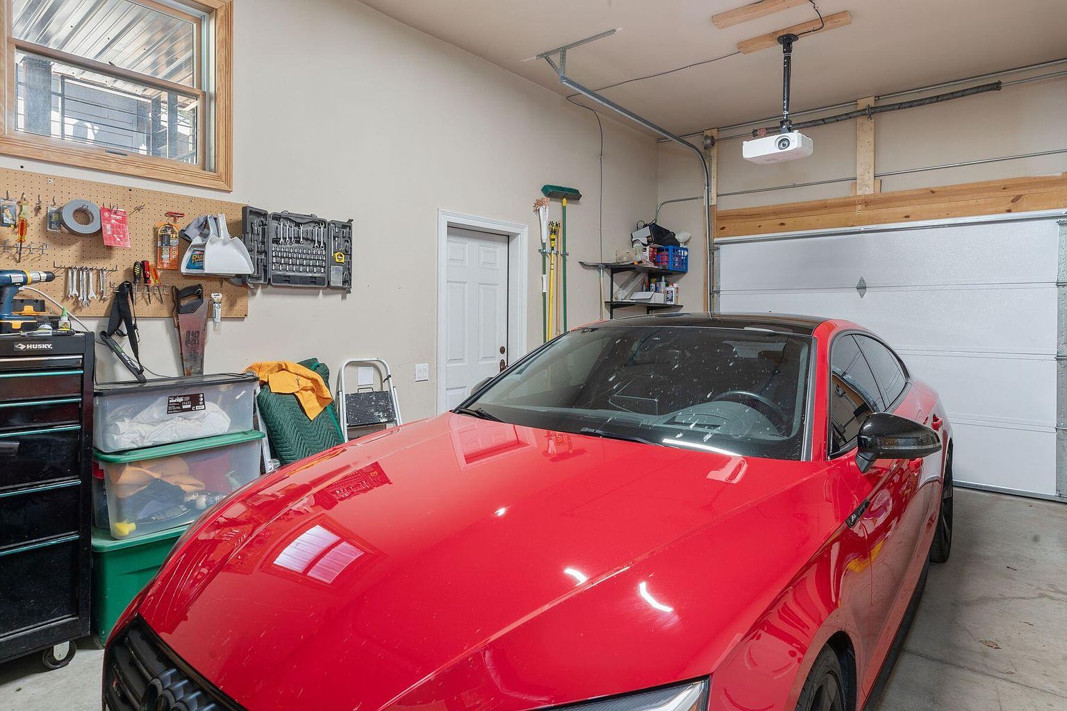 The image showcases a well-organized garage interior featuring a red car parked in the center. The garage includes storage solutions like toolboxes and plastic bins, along with a pegboard for hanging tools. A projector is mounted on the ceiling, suggesting a potential entertainment setup within the garage space.