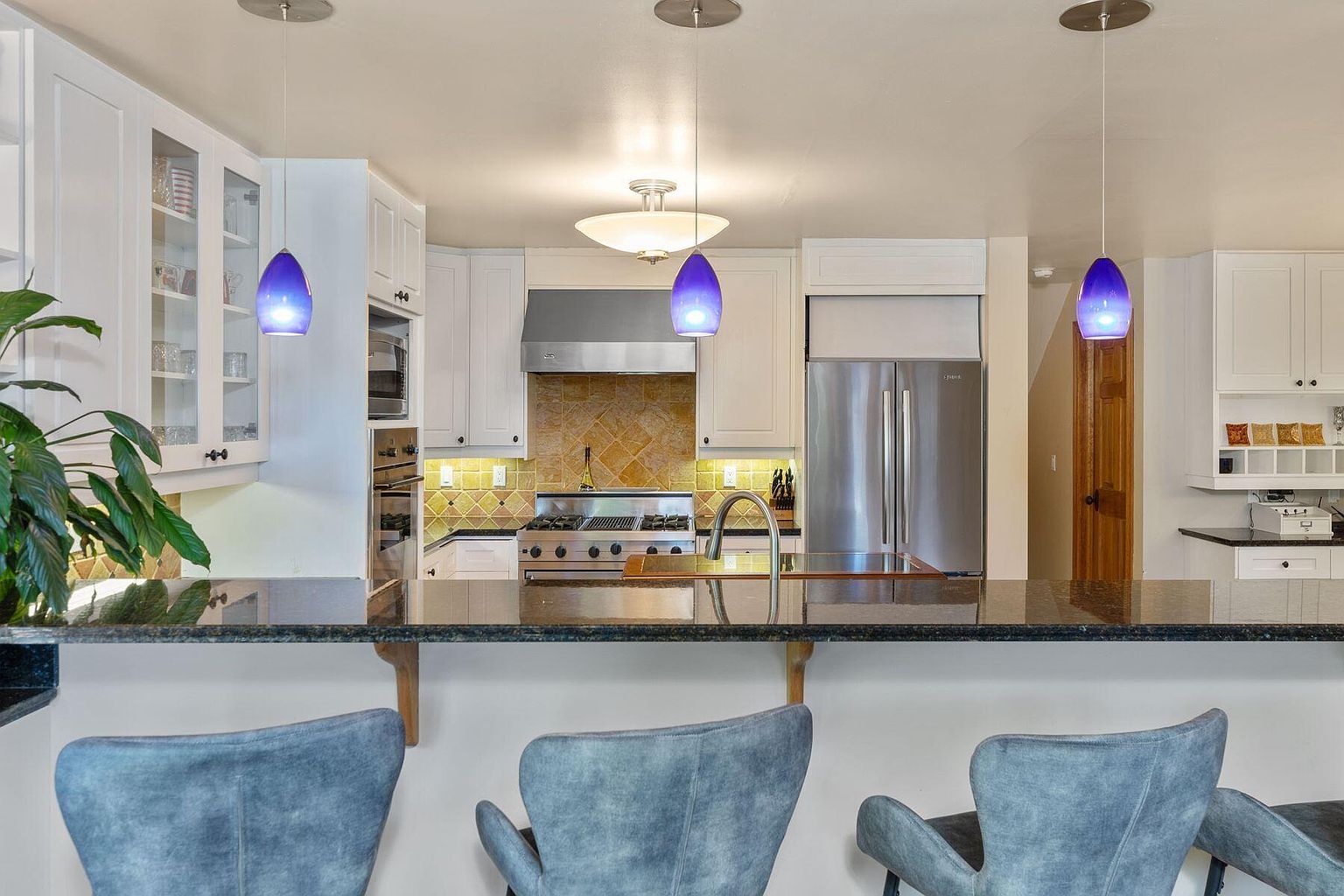 This is a well-lit kitchen featuring white cabinetry, stainless steel appliances, and a dark countertop with bar seating. Three blue pendant lights hang above the counter, adding a pop of color. The kitchen has a mix of modern and traditional elements, creating a warm and inviting atmosphere.
