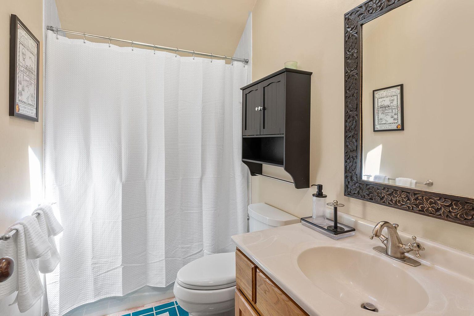 This is a well-lit bathroom featuring a white shower curtain, a toilet, and a vanity with a white countertop and wooden cabinets. A dark-colored medicine cabinet hangs above the toilet, and an ornate mirror is positioned above the sink. The overall impression is clean and functional, with a touch of traditional style.