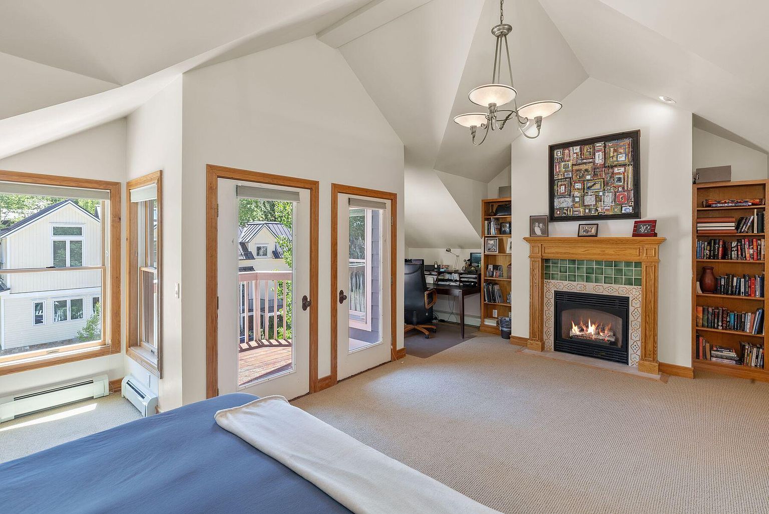 This is a primary bedroom featuring a fireplace with a decorative mantel and tile surround, flanked by built-in bookshelves. The room has a vaulted ceiling and includes a small office area with a desk and chair. Natural light floods the space through windows and a door leading to a balcony, creating a bright and inviting atmosphere.