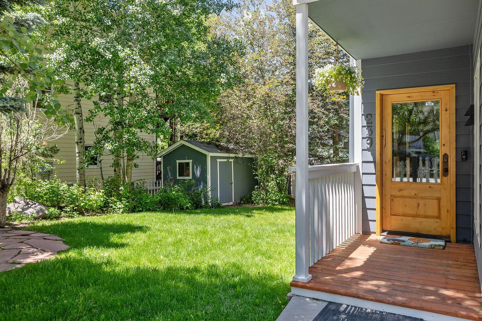 This image showcases a charming yard with a well-maintained lawn, mature trees, and a small shed in the background. The foreground features a wooden porch with a welcoming front door, suggesting a cozy and inviting home. The overall impression is one of tranquility and well-kept outdoor space.