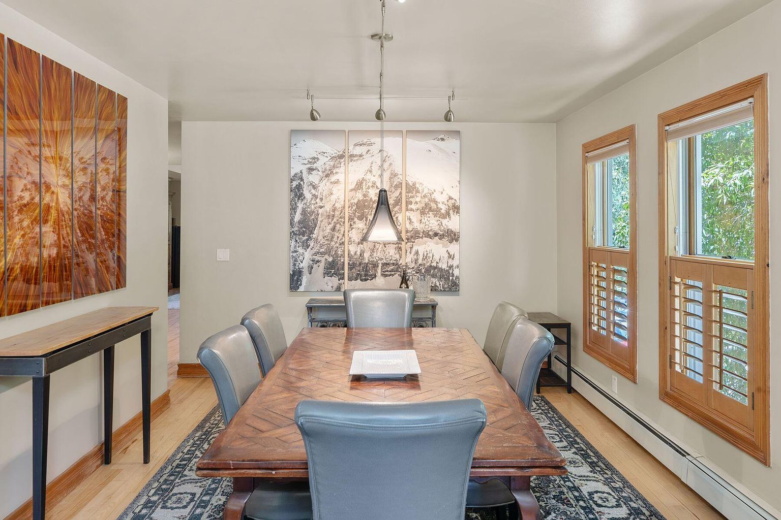 This is an interior shot of a dining room. The room features a large wooden dining table with gray leather chairs, a patterned rug, and two windows with wooden shutters. Artwork adorns the walls, and a modern light fixture hangs above the table, creating a warm and inviting atmosphere.